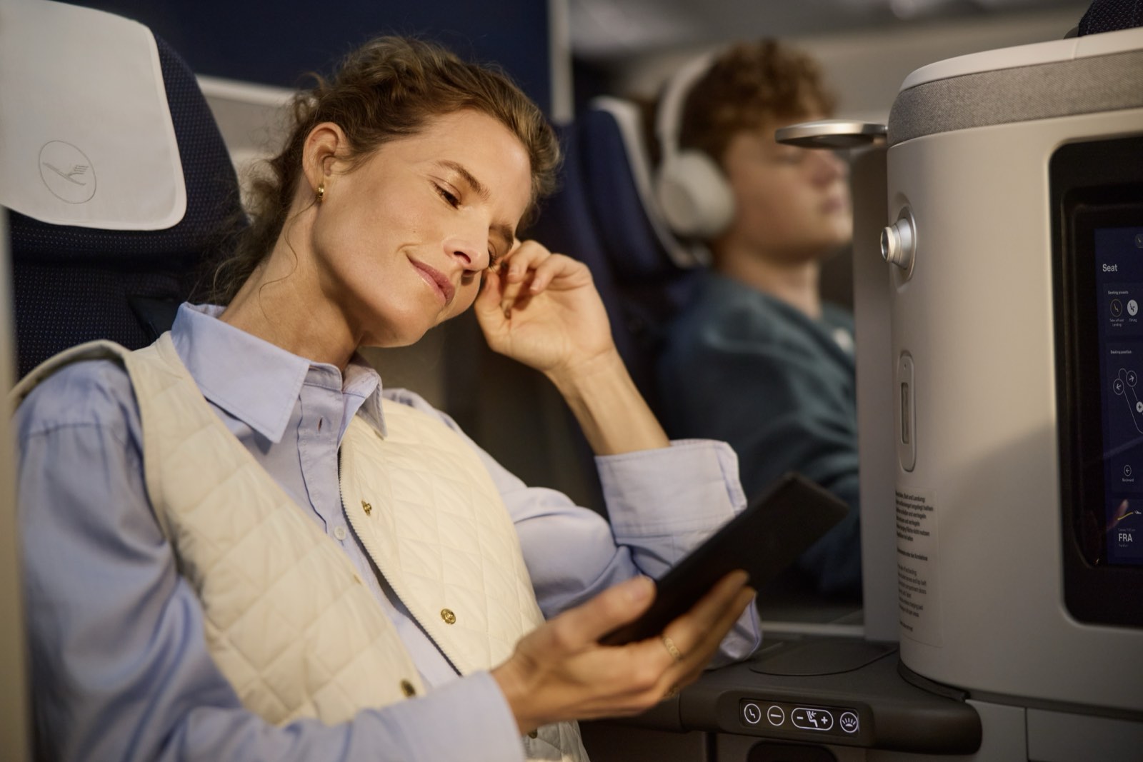 Ein Passagier in einem Lufthansa-Allegris-Business-Sitz mit Tablet, der Kranich auf der Kopfstütze und dahinter ein zweiter, zurückgelehnter Sitz